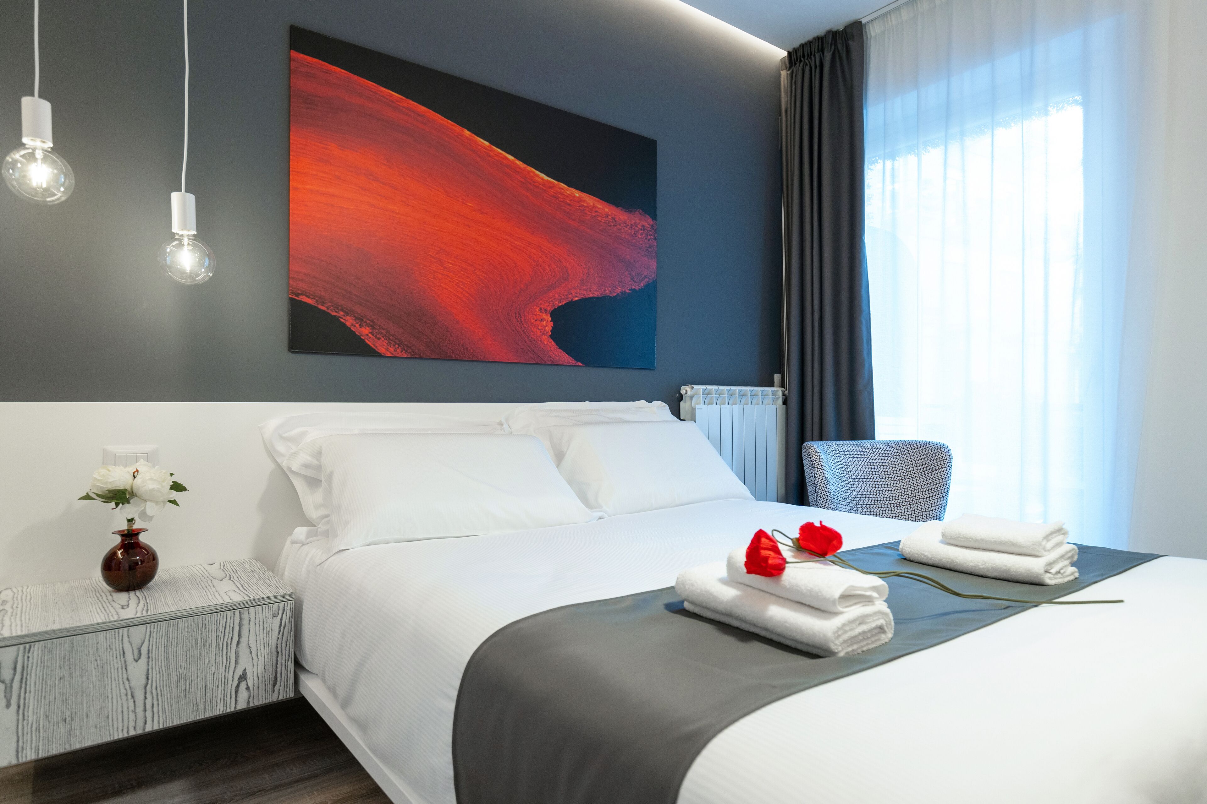 Una habitación de hotel moderna con una cama grande, una mesita de noche con un florero y una rosa roja sobre la cama, y un cuadro en la pared.