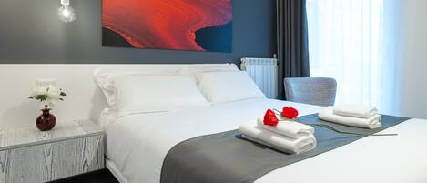 Una habitación de hotel moderna con una cama grande, una mesita de noche con un florero y una rosa roja sobre la cama, y un cuadro en la pared.