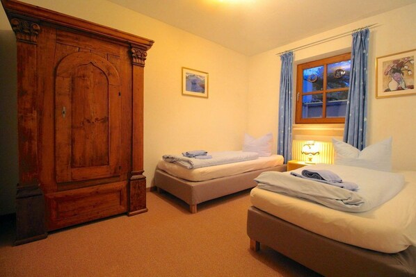 A hotel room with two single beds, a wooden wardrobe, a window with curtains, and a lamp on the nightstand.