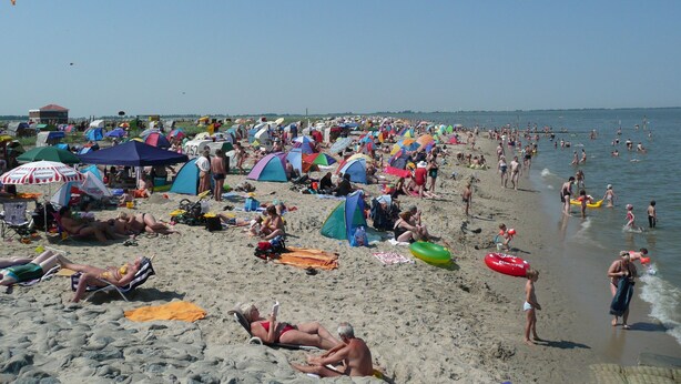 Ein überfüllter Strand mit vielen Zelten und Sonnenschirmen, Menschen, die sich sonnen und schwimmen, und einem klaren Himmel.