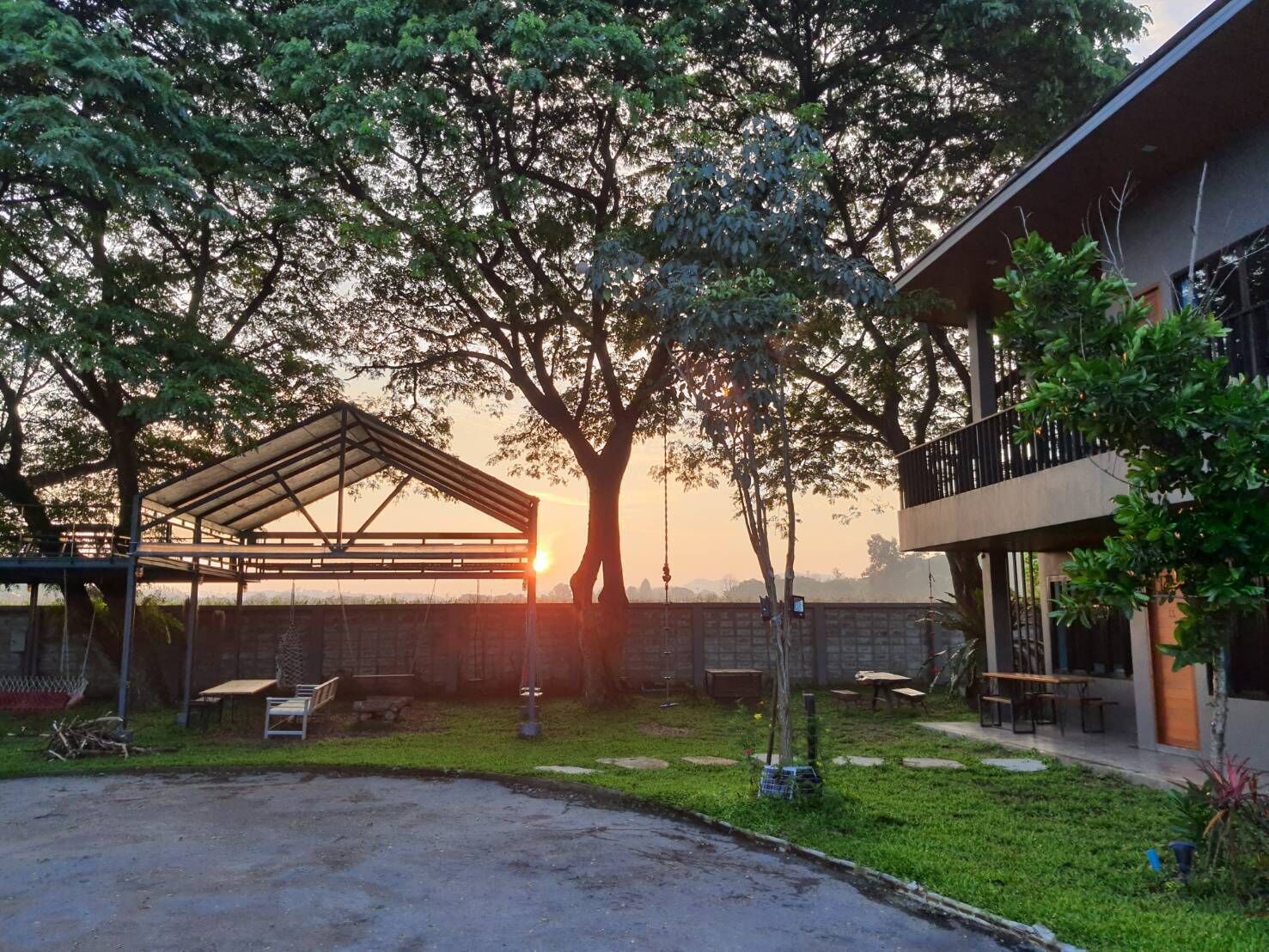 A covered outdoor area with a picnic table and a swing set, surrounded by trees and a building with a balcony.