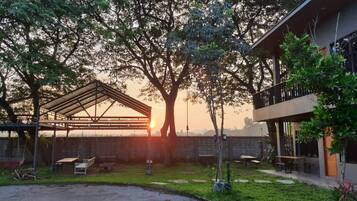 A covered outdoor area with a picnic table and a swing set, surrounded by trees and a building with a balcony.
