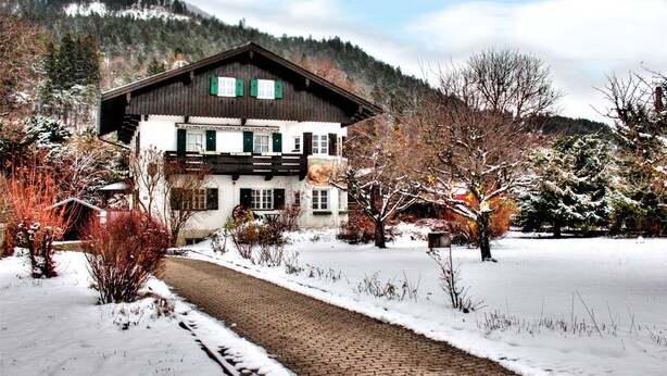 Ein traditionelles Chalet mit Holzhülle und grünen Fensterläden, umgeben von schneebedeckten Bäumen und blauem Himmel.