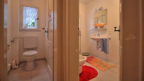 Ein Badezimmer mit Toilette, Waschbecken und Spiegel. Ein Fenster mit Vorhängen und ein roter Teppich liegen auf dem Boden.