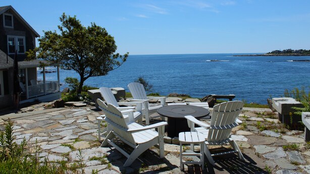 A stone patio with Adirondack chairs and a round table overlooking a body of water. - Ocean Front at Fortunes Rocks (Fortunes Rocks)