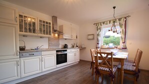 A kitchen with white cabinets, a wooden dining table with chairs, and a window with curtains. - Alpenschloss (Garmisch-Partenkirchen)