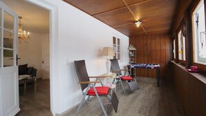 A room with wooden walls and ceiling, a sofa, two chairs with red cushions, a table with a lamp, and a window with a view. - Alpenschloss (Garmisch-Partenkirchen)
