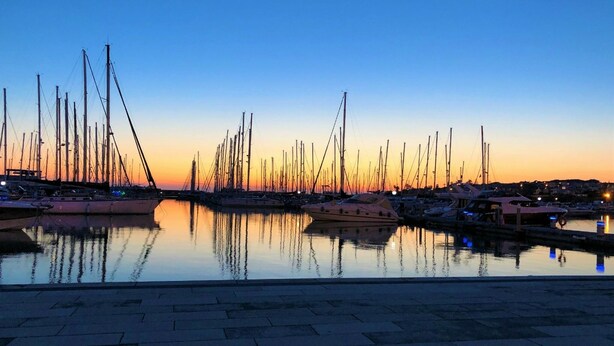 Un port de plaisance au coucher du soleil, avec de nombreux bateaux amarrés, des eaux calmes reflétant les bateaux et un ciel dégagé.