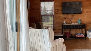 A cozy cabin interior with a wooden ceiling, a television mounted on the wall, a striped armchair, a small table with a vase, and a window with blinds. - Riverview Cottage on Green Gables Shore (Anglo-Rustico)