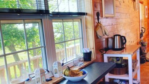A wooden cabin kitchen with a large window, a bowl of fruit, a kettle, and a coffee maker on the counter. - Riverview Cottage on Green Gables Shore (Anglo-Rustico)