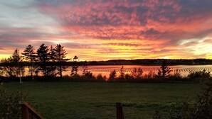 A sunset over a lake with a grassy area and wooden deck in the foreground. - Riverview Cottage on Green Gables Shore (Anglo-Rustico)