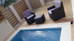 Ein kleiner Pool mit zwei Korbstühlen und einem Tisch, umgeben von einer Holzterrasse und einer weißen Mauer.