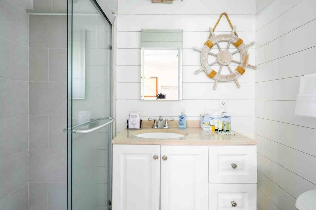 A bathroom with a glass shower enclosure, a white sink with a ship's wheel decoration, and a mirror above the sink.