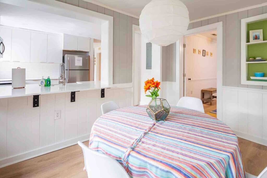 Una cocina moderna con armarios blancos, un refrigerador de acero inoxidable y un comedor con mantel a rayas de colores y un jarrón de flores naranjas.
