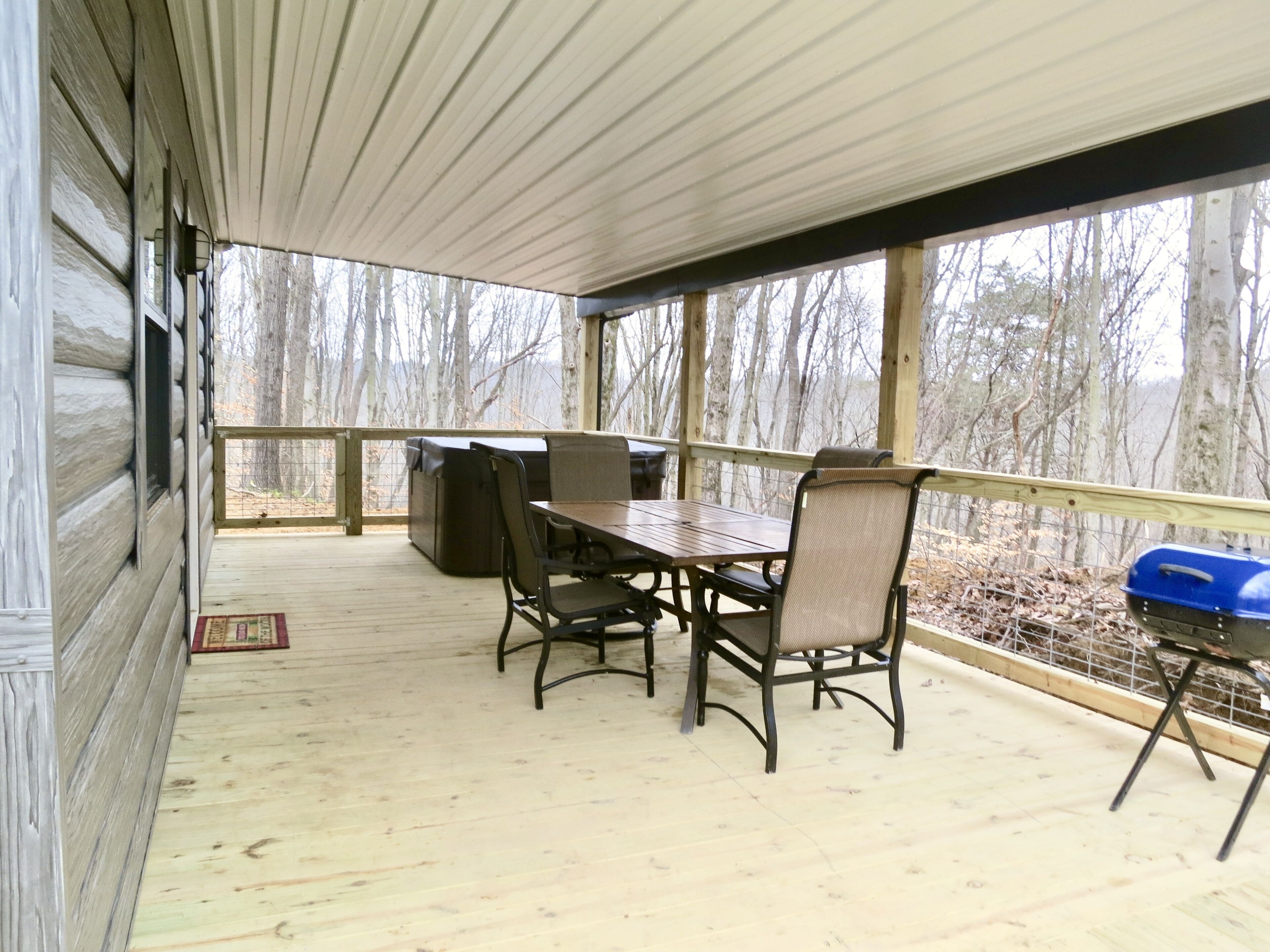 A covered deck with a wooden table and chairs, a grill, and a view of trees.