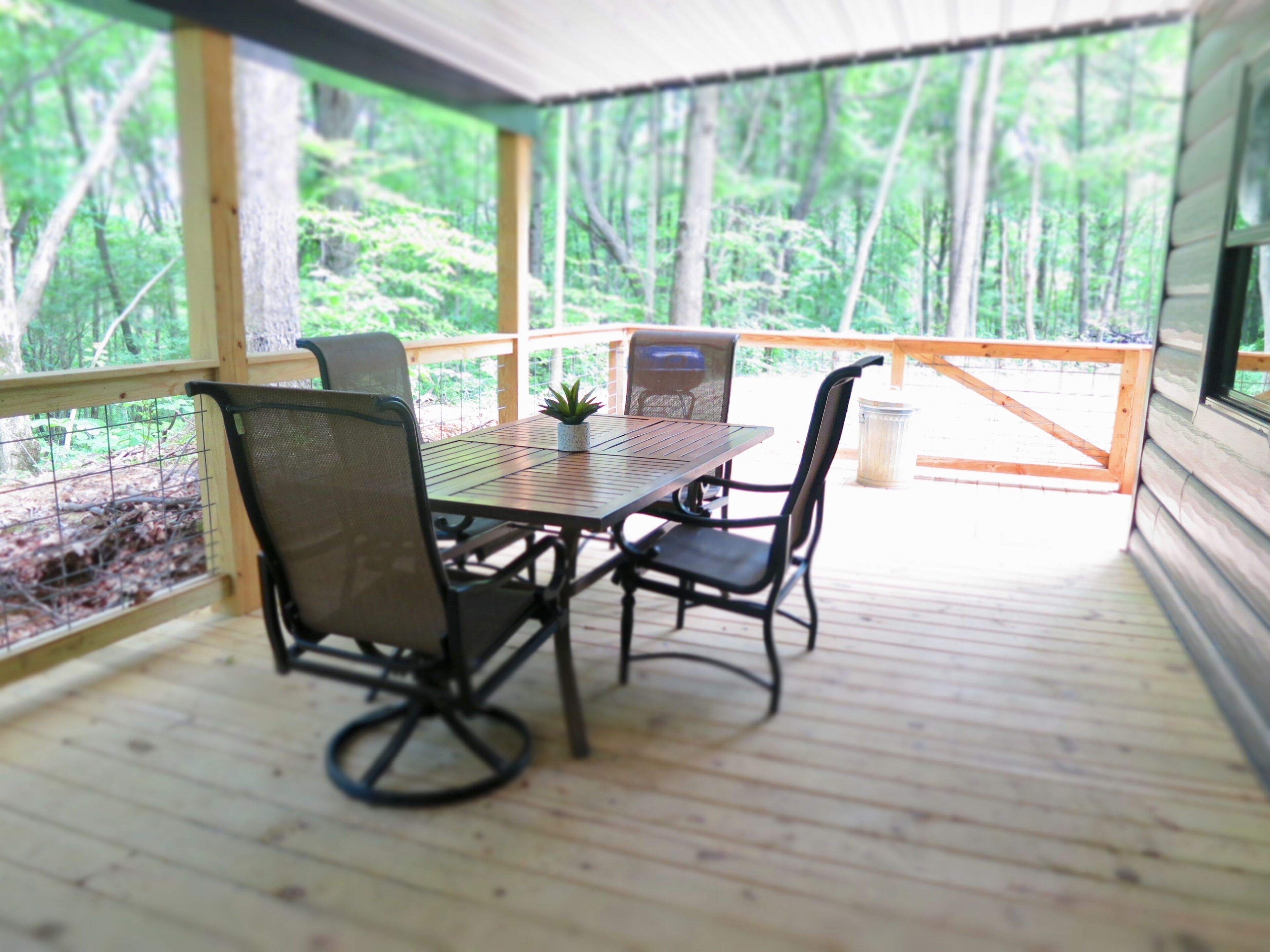 Uma área de refeições ao ar livre coberta, com mesa e cadeiras, cercada por um deck de madeira e vista para uma floresta.