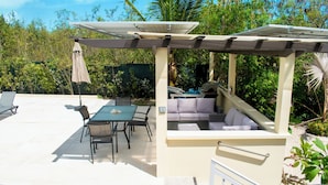 Un patio al aire libre con una mesa y sillas de comedor, una zona de estar con cojines y una pérgola con paneles solares.
