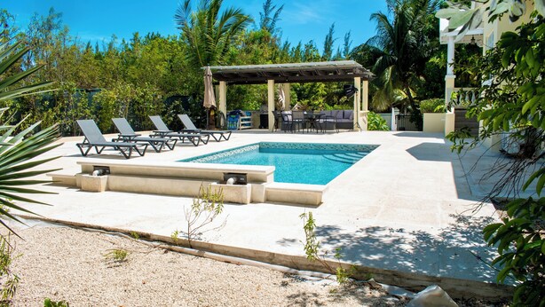 Un área de piscina con sillones reclinables, una parrilla y una zona de estar cubierta, todo rodeado de vegetación.