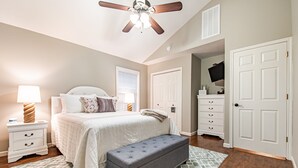 A bedroom with a bed, nightstand, bench, ceiling fan, and a door leading to another room.