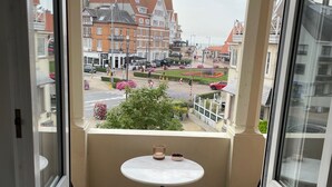 A balcony with a round table and two wooden benches, overlooking a street with buildings and flowers. - Charming renovated luxury apartment with seaview. (De Haan)