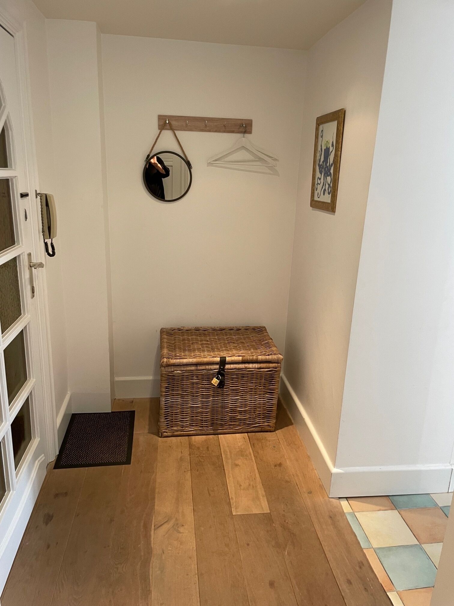 Una habitación de esquina con un cofre de mimbre, un perchero con espejo y un cuadro en la pared.