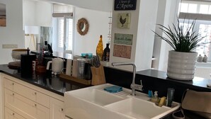 A modern kitchen with a large white sink, white cabinets, and a black countertop. There is a window with a view of the outside, and various kitchen items are visible on the counter. - Charming renovated luxury apartment with seaview. (De Haan)