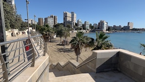 Un passeig marítim amb vista al perfil urbà de la ciutat, palmeres i una zona de platja de sorra.