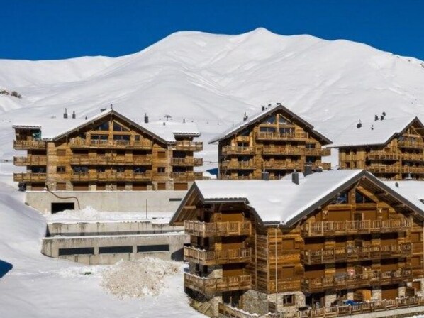 A cluster of wooden chalet-style buildings with balconies, set against a backdrop of snow-covered mountains.