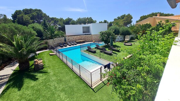 Una piscina rectangular con un deck perimetral, una pared de piedra y un edificio blanco al fondo.