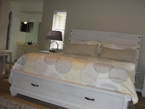 Quarto com cama, mesa de cabeceira, luminária, janela com persianas e espelho na parede.