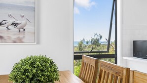 A dining area with a wooden table, chairs, a green plant, a framed picture of birds, and a view of the outdoors through a window. - Castaway Cove Castaways Beach (Castaways Beach)