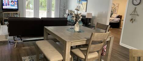 A dining area with a wooden table, chairs, and benches. There is a television in the background, a sofa, and a lamp on a side table.