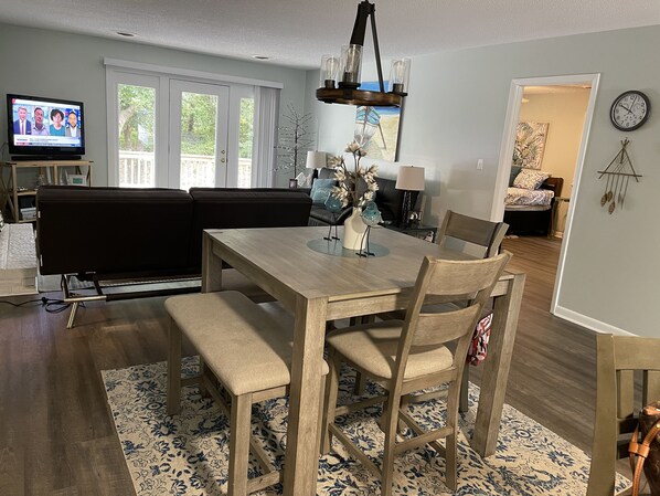 A dining area with a wooden table, chairs, and benches. There is a television in the background, a sofa, and a lamp on a side table. - Walk to beach / Cozy 3-Bedroom /Wrap-around deck (Pine Knoll Shores)