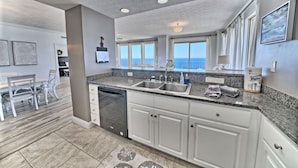 A modern kitchen with granite countertops, white cabinetry, and a dishwasher. There are large windows offering a view of the ocean.