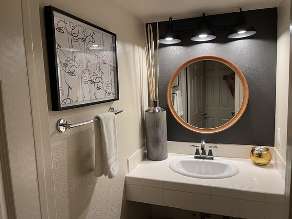 A bathroom with a white sink, a mirror with a wooden frame, a framed artwork, a towel rack, and a vase with reeds.