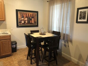 A dining area with a table set for four, surrounded by chairs. A framed painting of wine bottles hangs on the wall above the table.