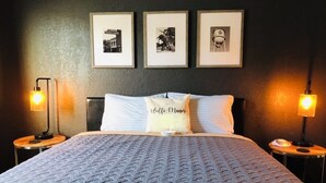 Un lit bien fait, recouvert d’une couverture matelassée, avec deux lampes de chevet et trois tableaux encadrés accrochés au mur.