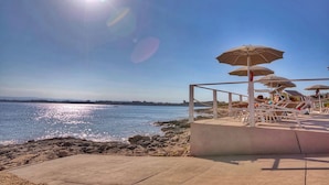 A beachside area with lounge chairs and umbrellas, a concrete walkway, and a rocky shoreline. - ARTEMIDE HOUSE - Superior Apartment Ortigia Wifi (Siracusa)