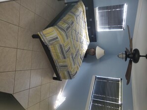 Relax in this serene space with a comfortable bed against a window with horizontal blinds, a ceiling fan above and a lamp nearby.