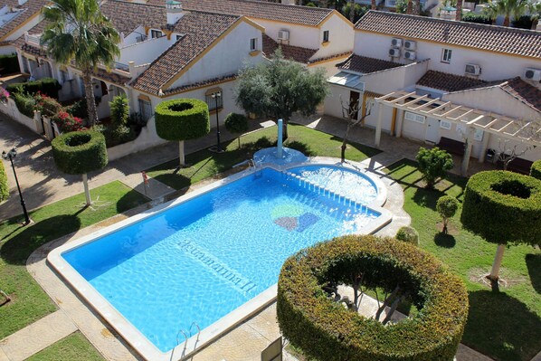 Una piscina rodeada de setos cuidados y una pequeña fuente, con edificios residenciales al fondo.