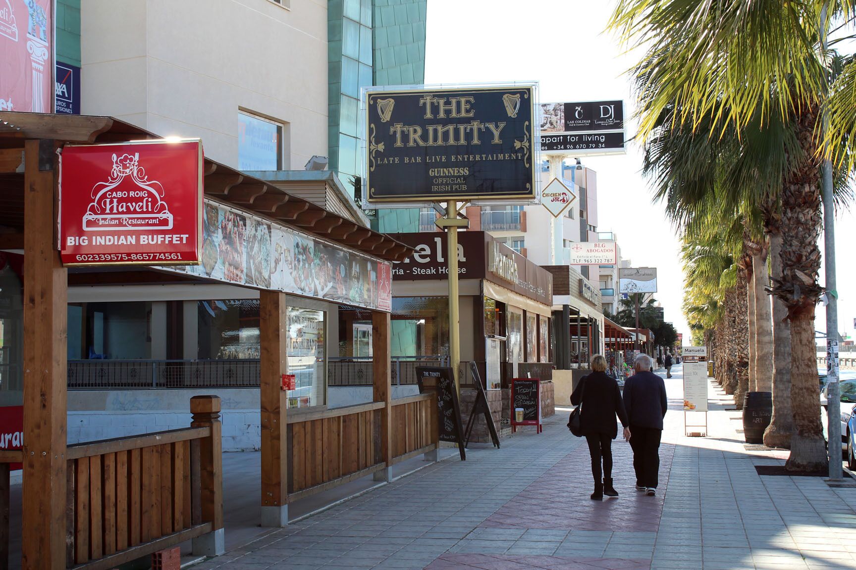 Une rue bordée de divers commerces et restaurants, dont on peut voir les enseignes de « The Trinity » et de « Cabot Roig Havelis ».