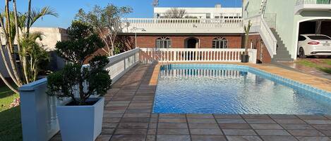Una piscina con borde de baldosas, rodeada por una valla blanca y macetas con plantas.
