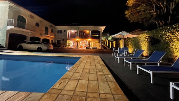 A pool area with lounge chairs and umbrellas, a building with multiple levels and a covered patio, and a car parked nearby. - Dom Ravello (Telemaco Borba)