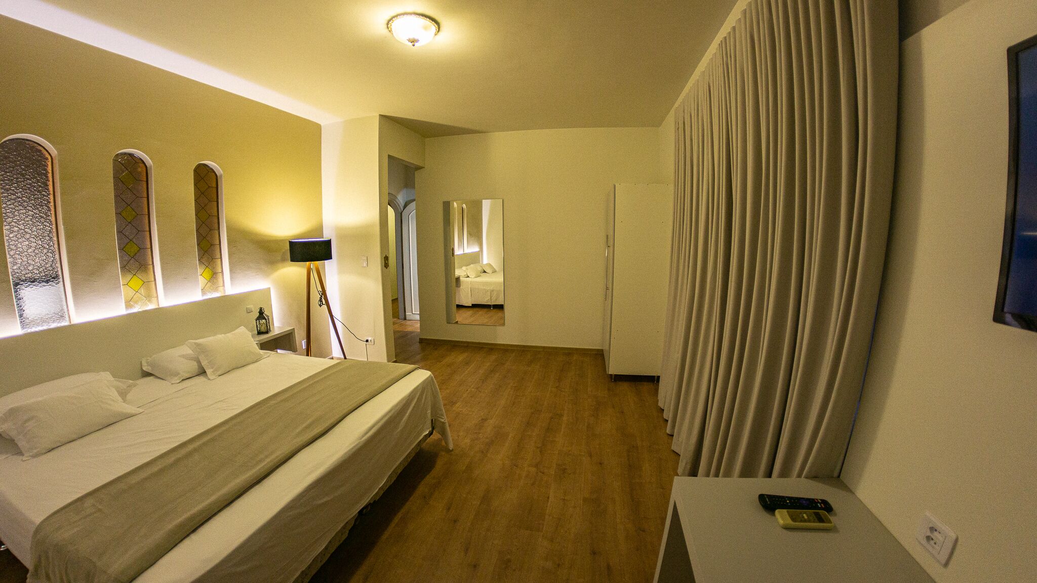 Habitación de hotel con cama, mesita de noche con control remoto, lámpara de pie y espejo en la pared.