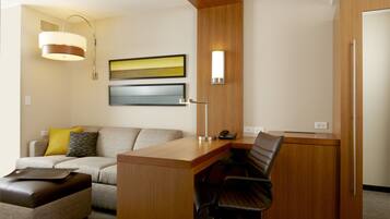 Habitación de hotel con un escritorio de madera, una silla de cuero, un sofá con cojines, una lámpara y dos cuadros en la pared.
