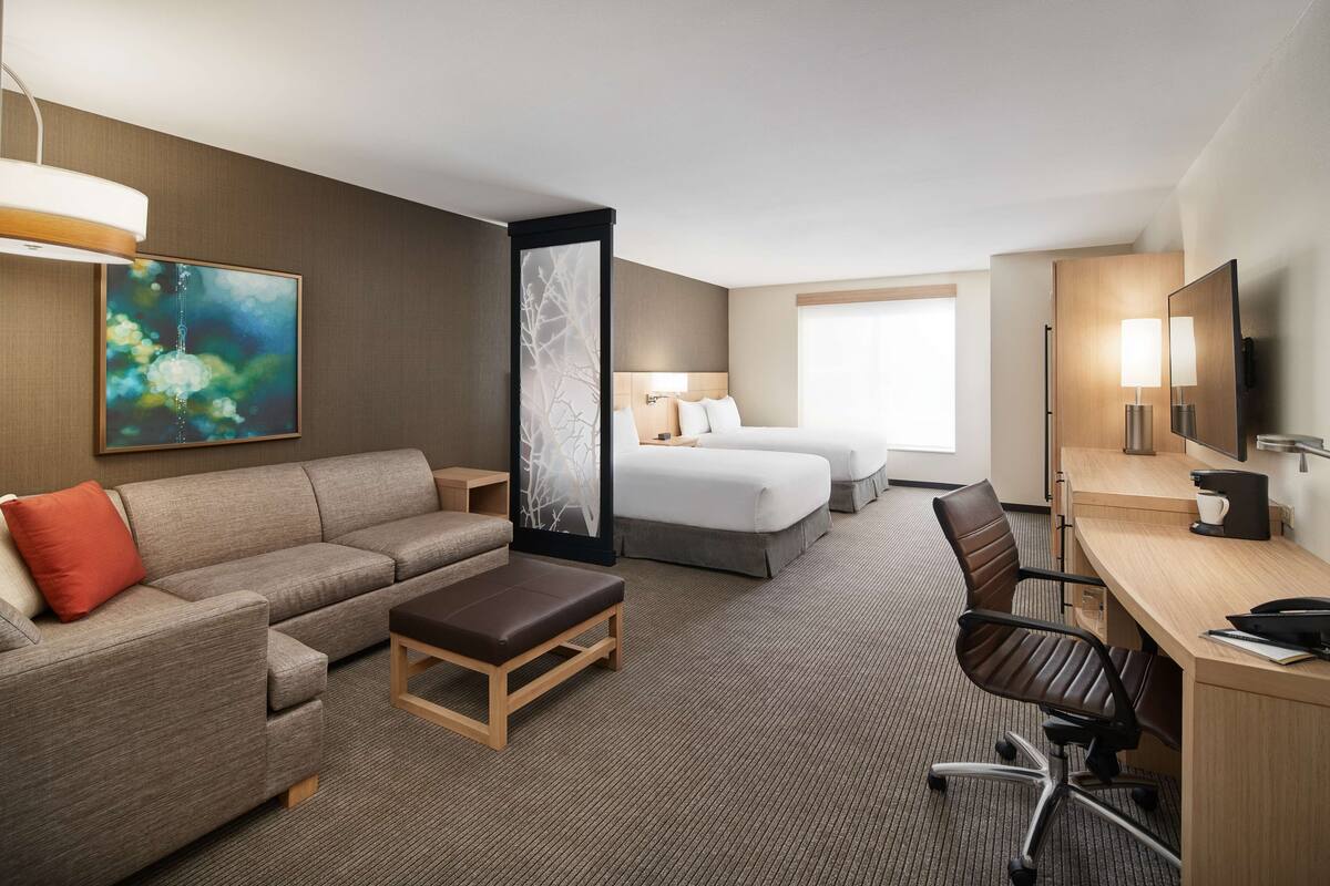A hotel room with a sofa, two beds, a desk, and a large window.