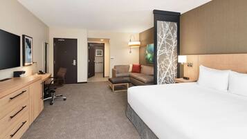 A hotel room with a large bed, a desk with drawers, a chair, a sofa, a TV, and a door leading to another room.