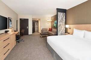 A hotel room with a large bed, a desk with drawers, a chair, a sofa, a TV, and a door leading to another room.
