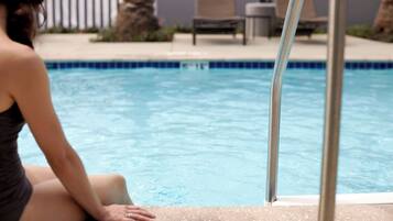 Une personne assise au bord d’une piscine, avec des chaises longues et des palmiers en arrière-plan.