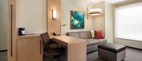 Una habitación de hotel moderna con un escritorio de madera, un sofá gris, un reposapiés marrón y un ventanal grande.